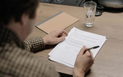 A businessman is reviewing a contract while holding a pen at a desk with a glass of water nearby.