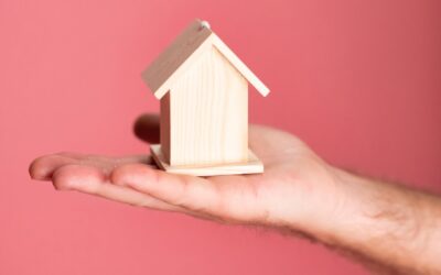 Wooden house model in hand symbolizing real estate concepts on pink background.